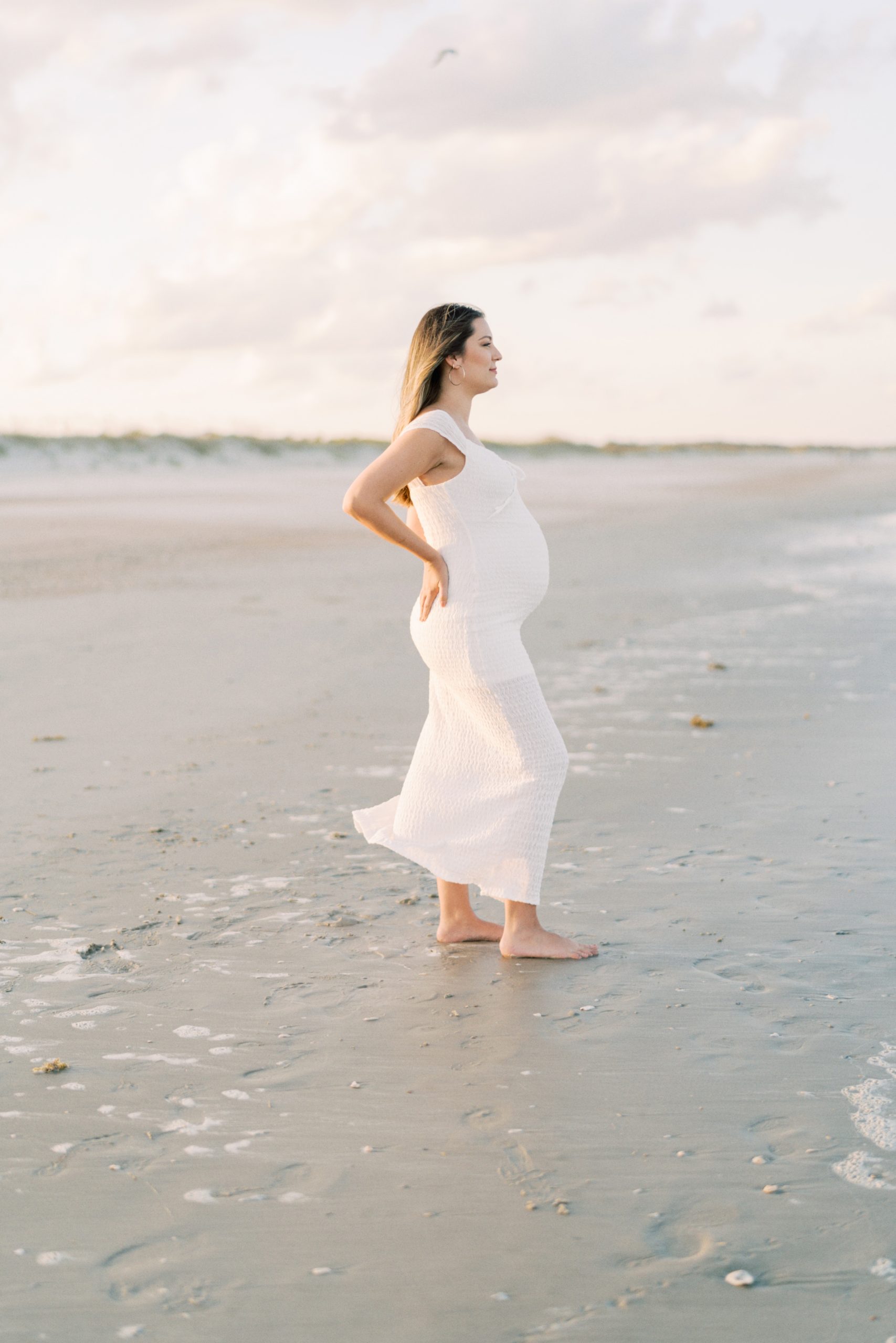 Pregnant woman holding belly and looking out to the beach during her maternity session.