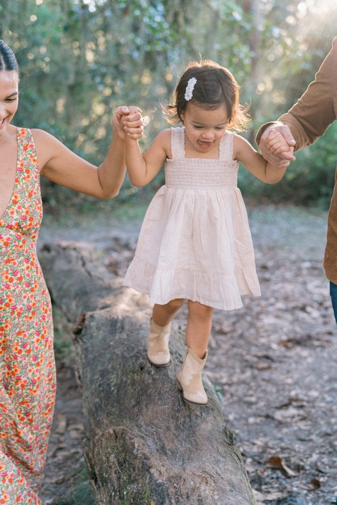 Parents holding daughters hand while she walks on a tree branch.