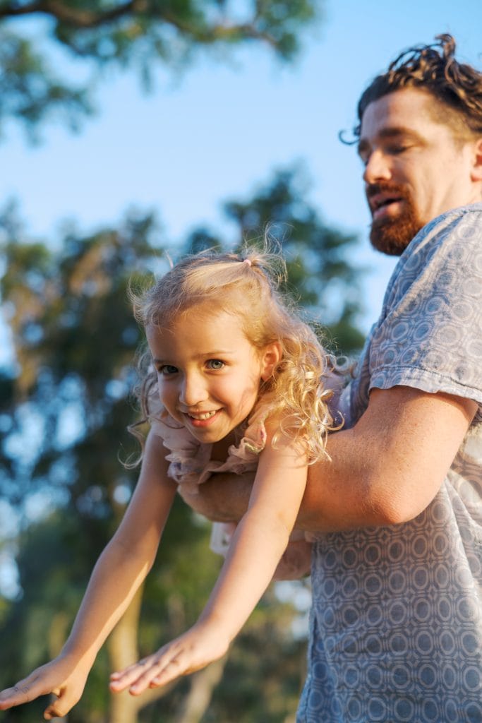 Father lifting young girl with curly blonde hair, smiling happily in a park setting.